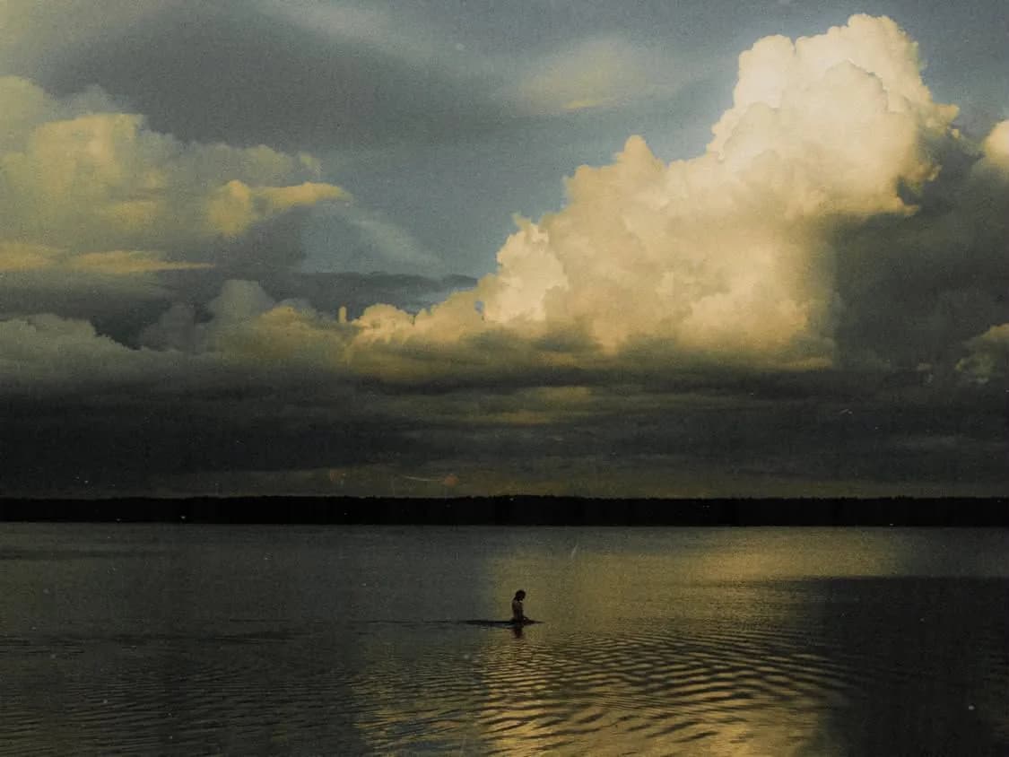 Lone swimmer in a calm lake under towering clouds